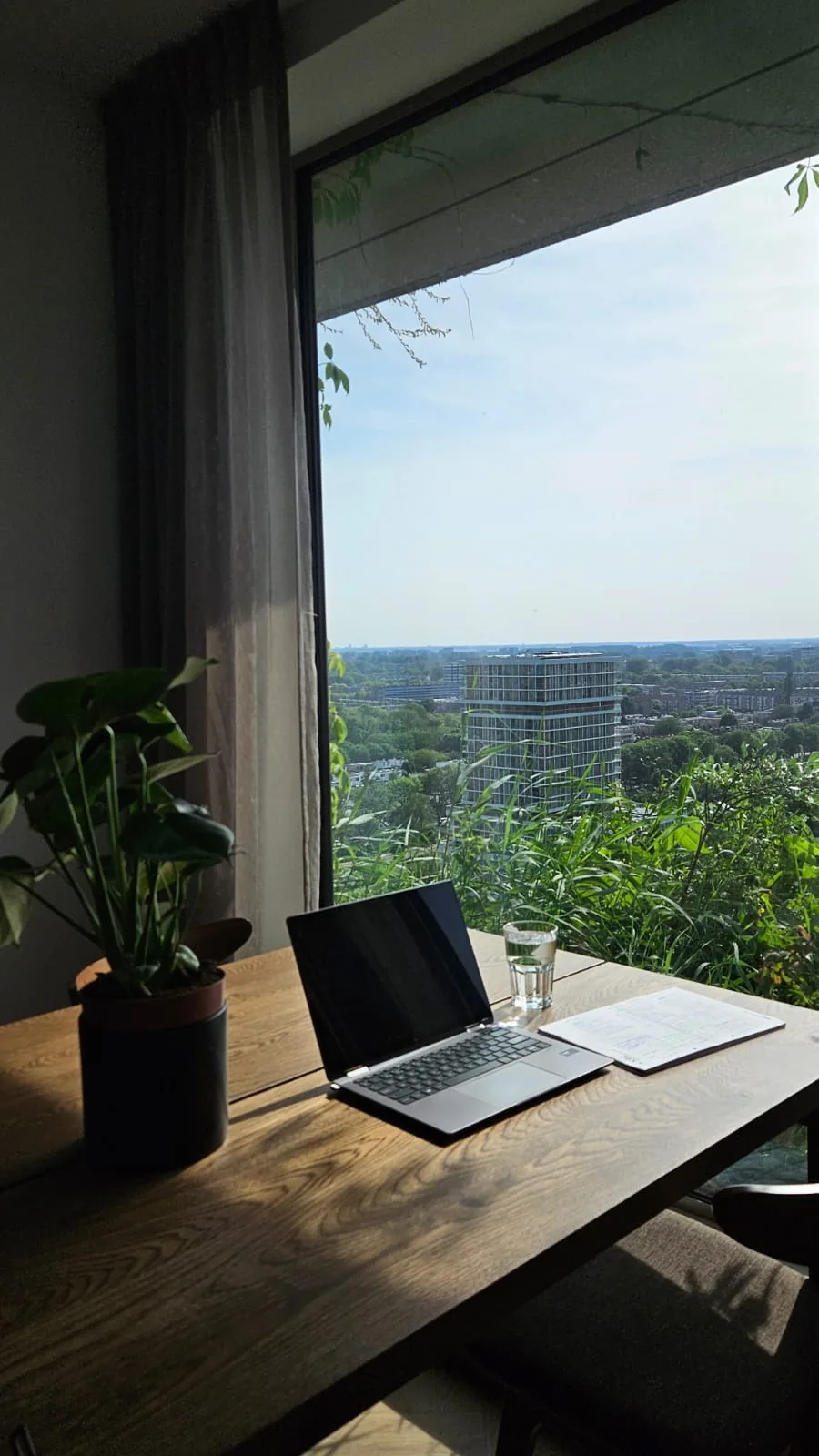 Laptop auf einem Holztisch vor grosser Fensterfront mit Blick über eine grüne Stadtlandschaft